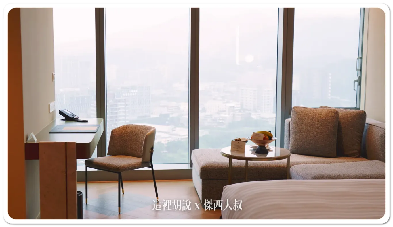 The room's got these big floor-to-ceiling windows with both blackout curtains and sheer curtains. So you can choose how much light or privacy you want. They've set up a desk and chair by the window, along with a comfy sofa. It's a great spot to work or just chill out with a view.