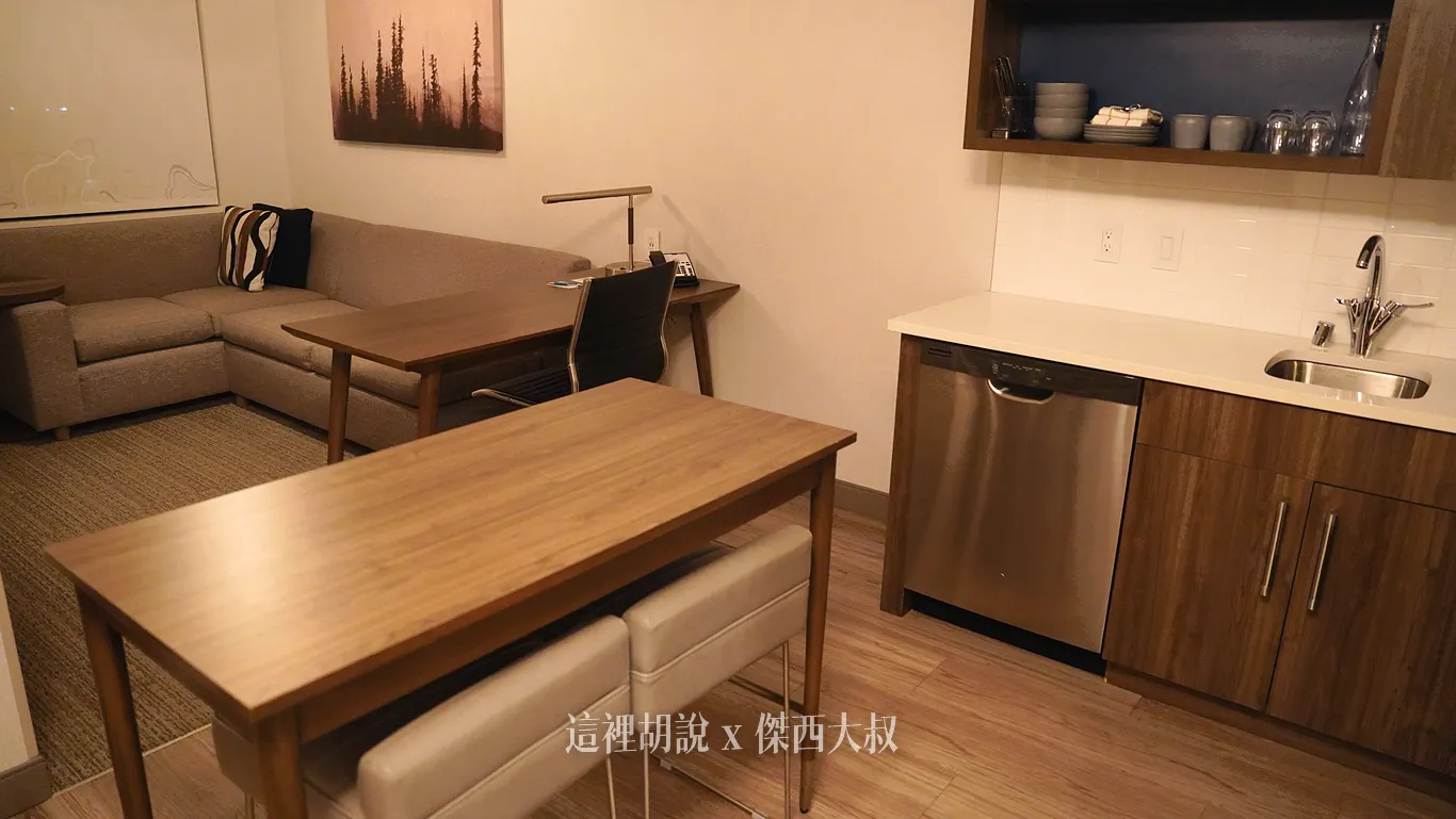 除了後方的沙發與辦公書桌，前方木質地板上還設有一座搭配兩張高腳椅的木質餐桌用餐空間，右側則銜接配備水槽與洗碗機的廚房吧檯。