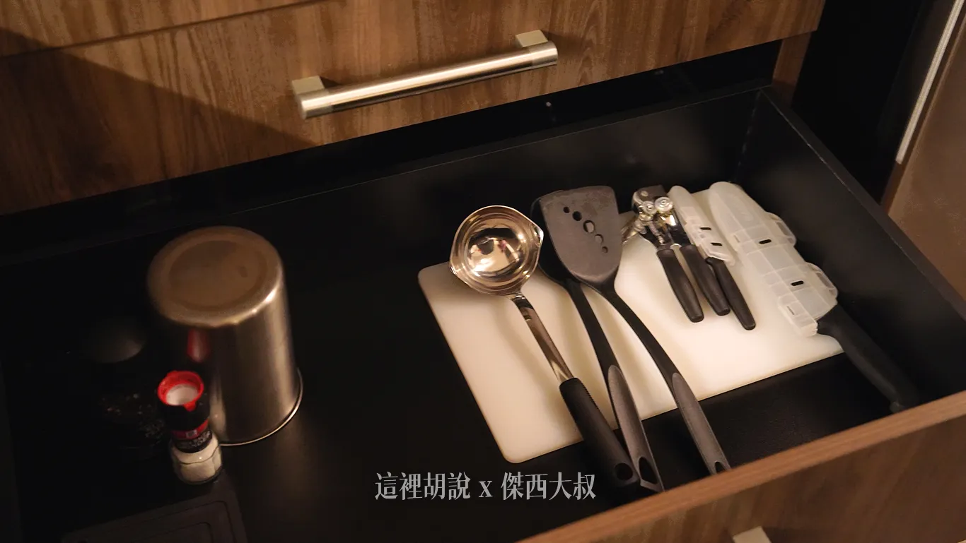 廚房抽屜內整齊收納調理用的金屬湯勺與鍋鏟，一旁白色砧板上還備有開罐器、帶保護套的刀具以及調味罐等料理用具。