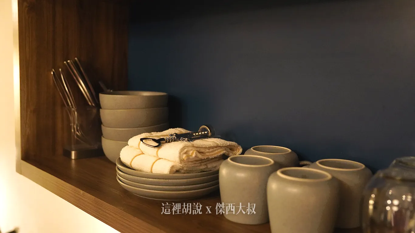 西雅圖機場Element Hotel廚房木質層架上擺設整齊齊全的餐具，包含疊放的灰色陶瓷碗盤、馬克杯與玻璃杯，旁邊備有折疊抹布、開瓶器及刀叉餐具筒。