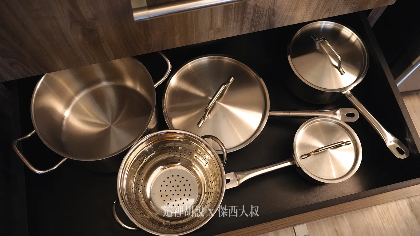 抽屜內，整齊擺放著雙耳湯鍋、瀝水篩網盆、平底鍋與不同尺寸的單柄帶蓋不鏽鋼鍋具，呈現乾淨明亮的光澤。