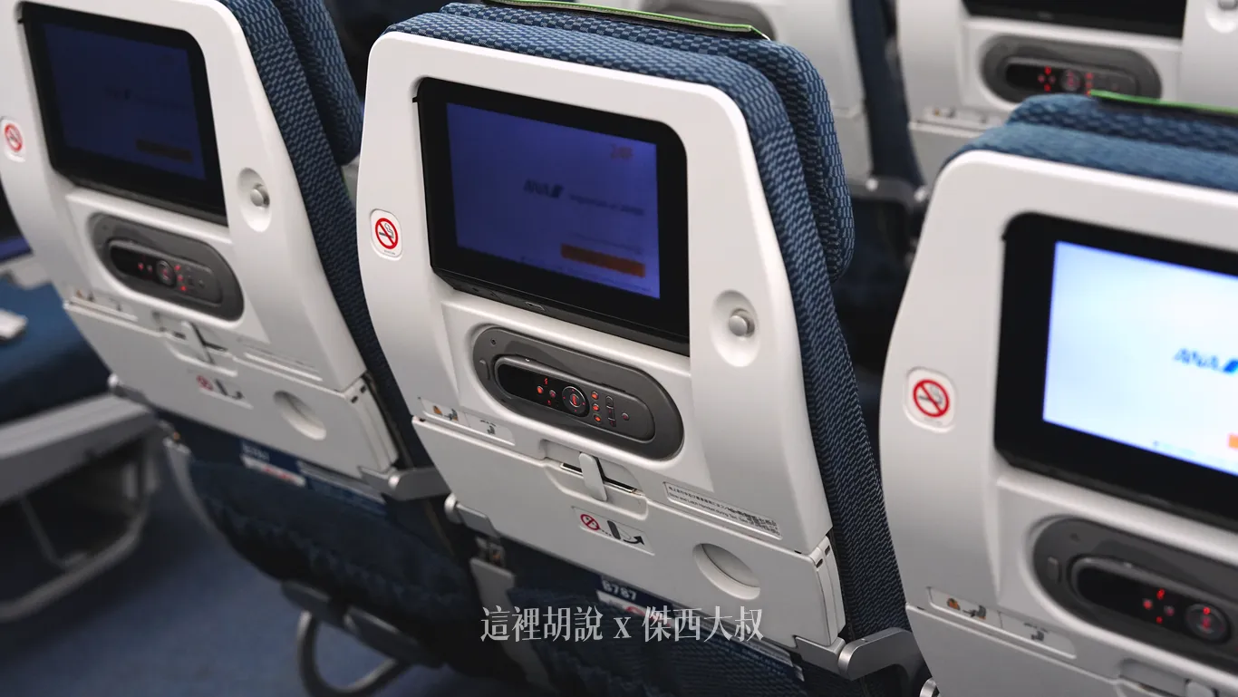 全日空787-9客艙經濟艙座椅椅背嵌有個人娛樂系統螢幕與專用遙控器，下方設有附杯架的兩段式折疊桌板，白色背板搭配深藍色織布椅套。