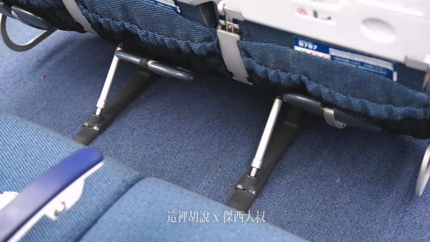 全日空787-9客機經濟艙座椅下方設有金屬支架的可折疊腳踏板，藍色織布椅背下方配有分層的多功能收納網袋，提供乘客放置隨身物品與長途航程的置腳空間。