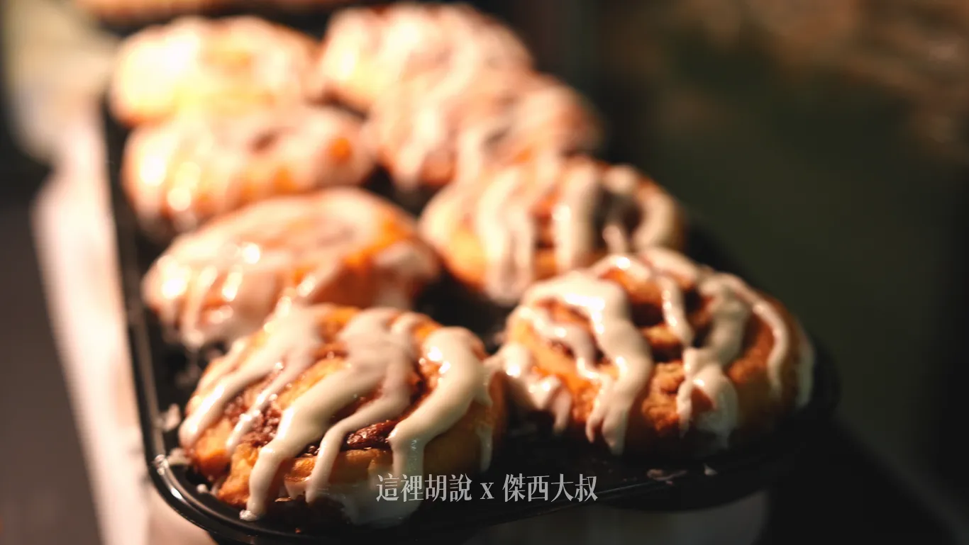 肉桂捲整齊排在黑色托盤上,烘烤至金黃酥脆的螺旋麵包頂端淋滿濃稠微融的白色糖霜,散發誘人香氣。 肉桂捲整齊排在黑色托盤上,烘烤至金黃酥脆的螺旋麵包頂端淋滿濃稠微融的白色糖霜,散發誘人香氣。