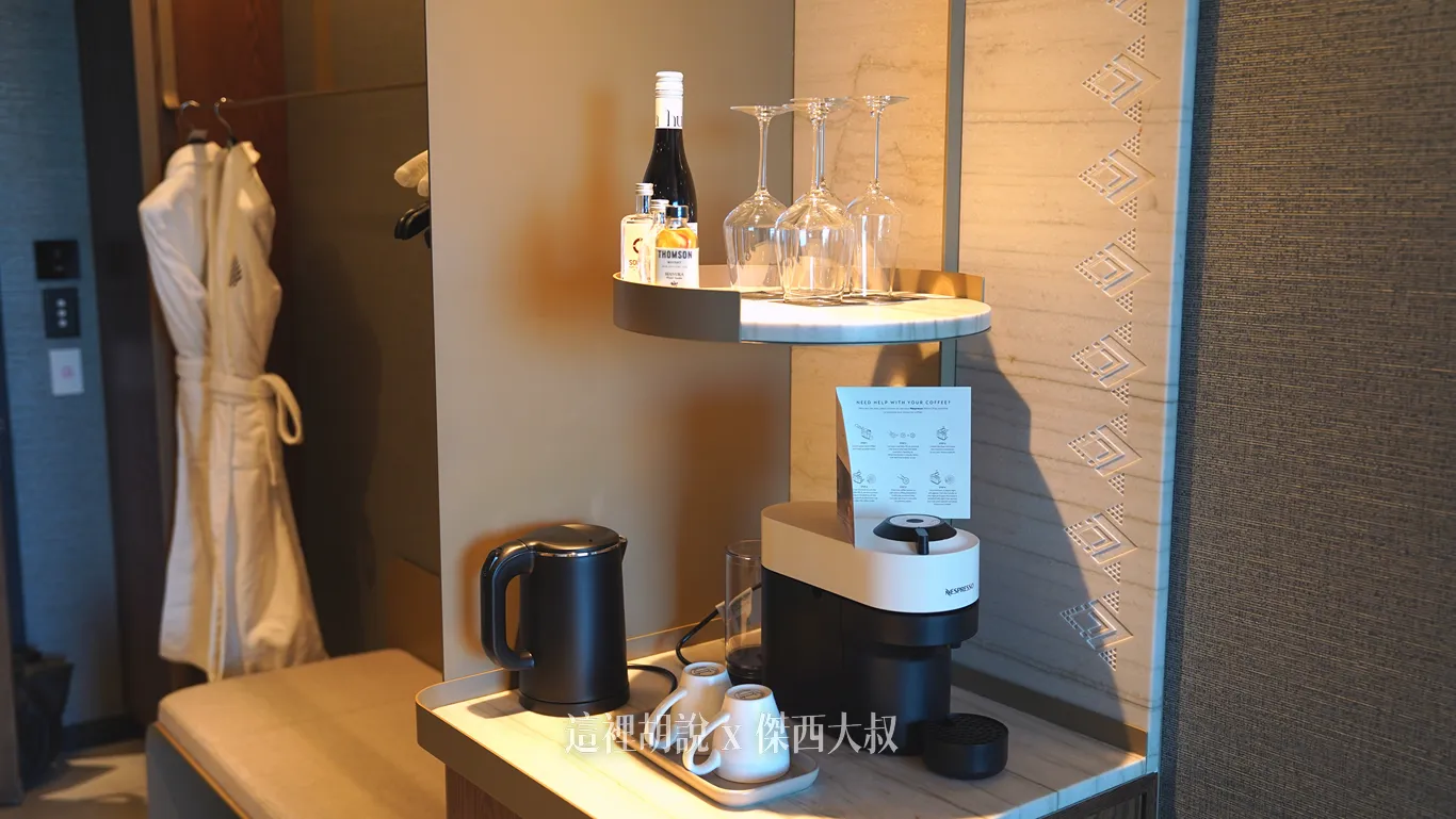 奧克蘭機場鉑爾曼飯店客房的迷你吧檯面上擺放黑色快煮壺、膠囊咖啡機與白色咖啡杯，上方圓形層架則備有酒杯與瓶裝酒品，一旁牆面飾有圖騰。