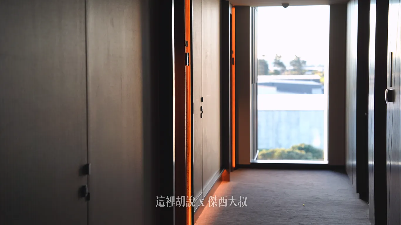 奧克蘭機場鉑爾曼飯店的客房外走廊鋪設灰色厚地毯，兩側深色木質牆面嵌有垂直的暖橘色間接照明燈條，盡頭設有大片採光落地窗並可見戶外景致。