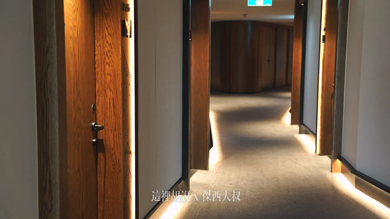 奧克蘭機場鉑爾曼飯店客房走廊呈現大氣簡約的設計風格，兩側為木質門扉與白色牆面，地面鋪設灰色地毯，牆腳內嵌暖黃色間接照明營造沉穩動線。