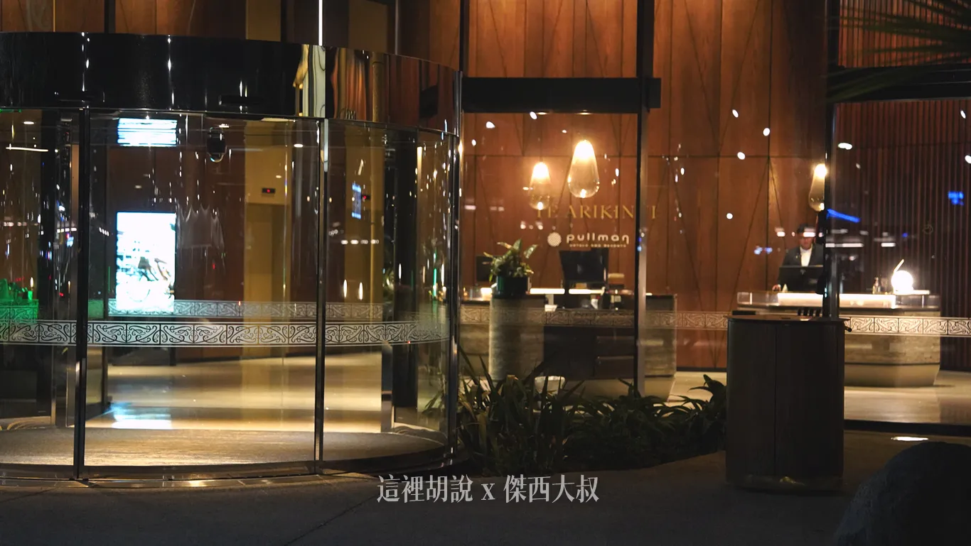 奧克蘭機場鉑爾曼飯店入口旋轉門玻璃飾有精緻圖騰腰帶，透過落地玻璃可見木質牆面、接待櫃檯與暖黃吊燈，營造沉穩雅致的大廳氛圍。