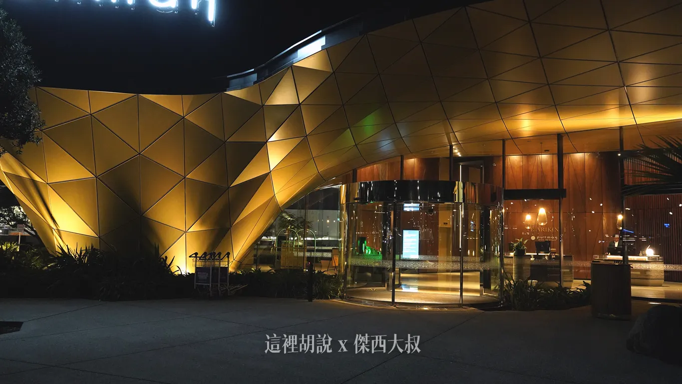 奧克蘭機場鉑爾曼飯店晚間的入口處，幾何金屬切面造型的遮雨棚在投射燈照射下散發溫暖金光，透明旋轉門映著大廳櫃檯的柔和照明。