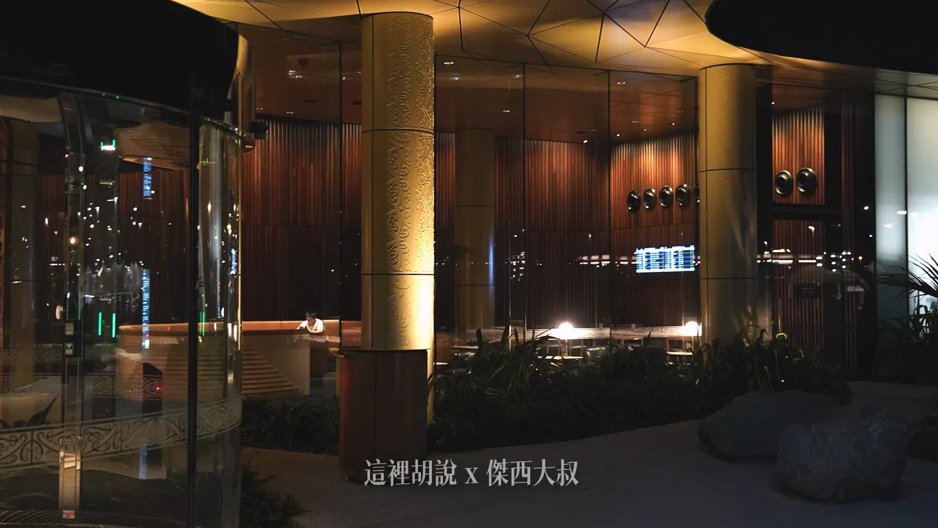 奧克蘭機場鉑爾曼飯店客房一側的休憩區，圓柱雕刻著細緻雕花圖騰，隔著大片落地玻璃映出木質格柵背景、圓弧形沙發與溫暖的投射燈光。