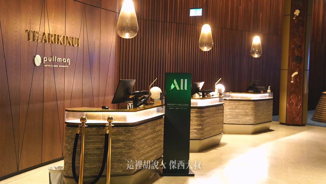 奧克蘭機場鉑爾曼飯店的大廳接待櫃檯，木質護牆板上標有金色品牌名稱，石紋曲面櫃檯上方垂掛著水滴狀玻璃吊燈，前方並設有雅高心悅界的會員優先報到立牌。