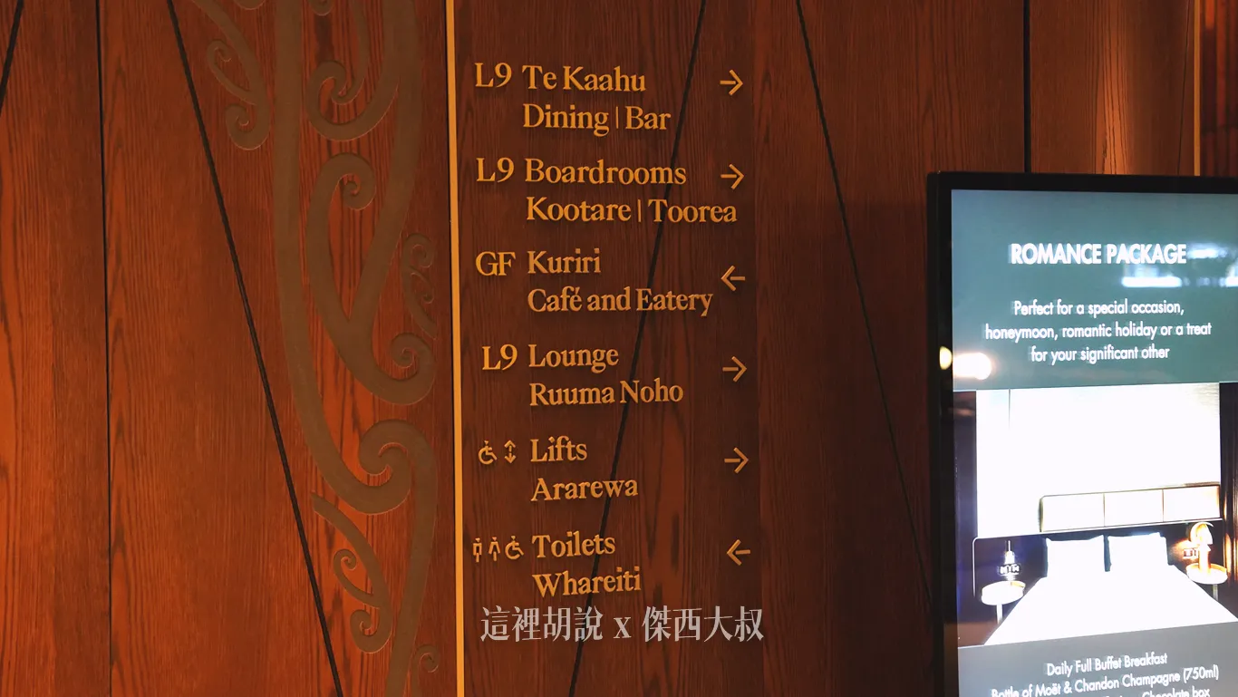 奧克蘭機場鉑爾曼飯店深色木質牆面上設有指標，除了英文之外同時並列毛利文樓層標示，旁邊還有一面播放客房專案資訊的直立式螢幕。