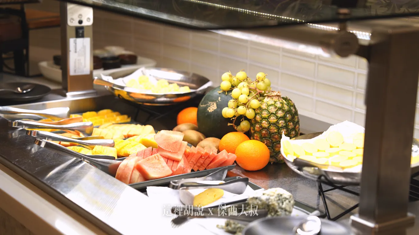 奧克蘭機場鉑爾曼飯店早餐自助吧台，陳列切片西瓜、切塊鳳梨與柳橙等新鮮水果，後方點綴整顆鳳梨、西瓜與葡萄裝飾，並備有取餐夾方便夾取。