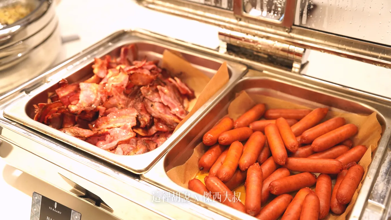 奧克蘭機場鉑爾曼飯店早餐自助餐檯上，金屬保溫餐爐內襯著烘焙紙，左右分別盛裝著煎至微焦的培根片與色澤紅潤飽滿的香腸。