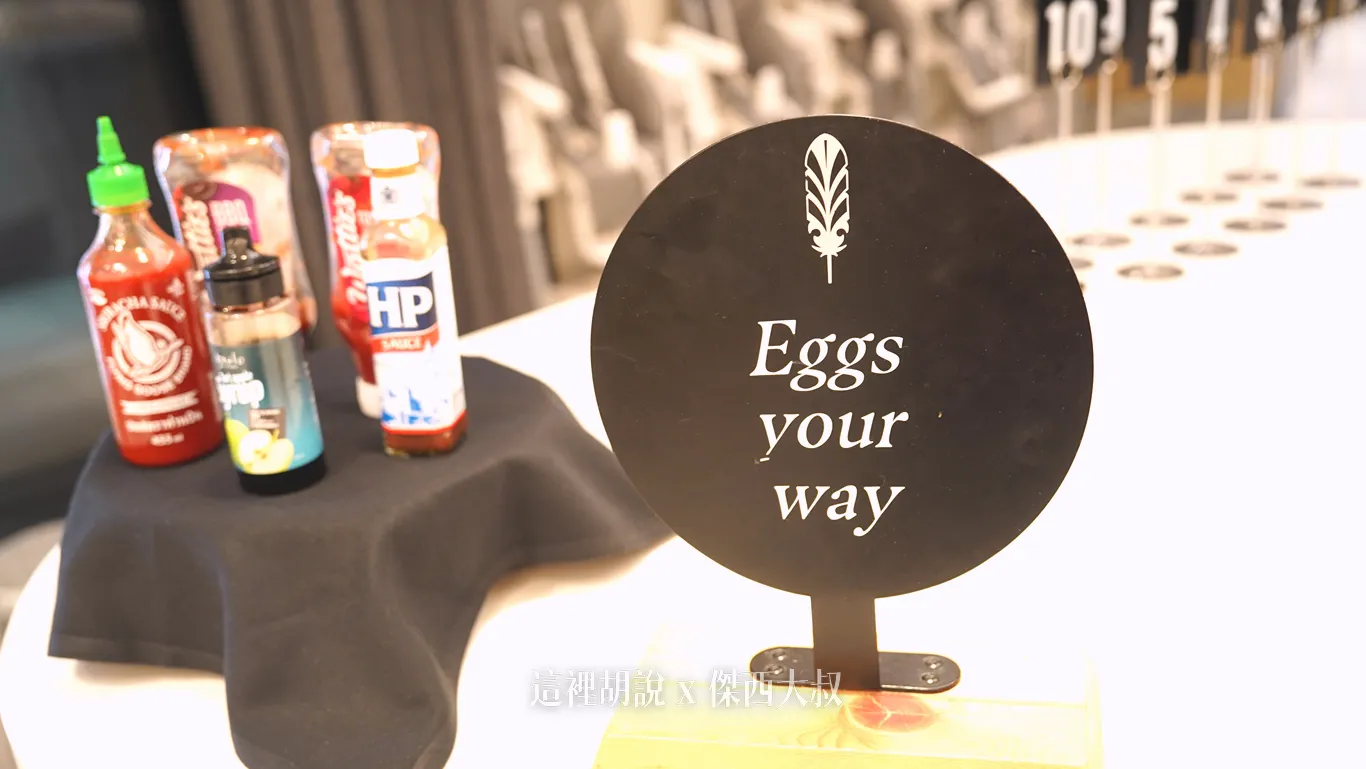 奧克蘭機場鉑爾曼飯店早餐吧檯立著寫有Eggs your way的黑色圓形告示牌，提供客製化煎蛋服務，旁側陳列著辣椒醬與HP醬等各式調味醬料供取用。
