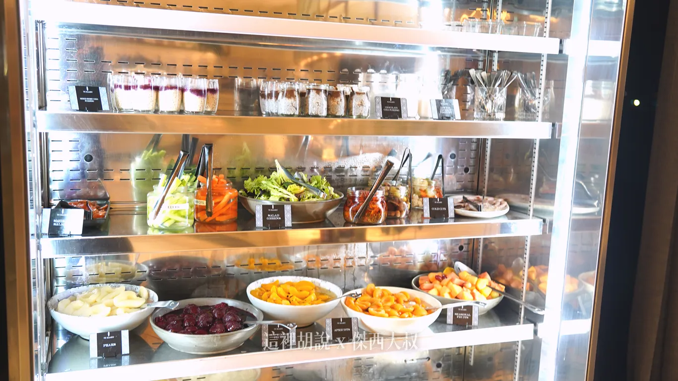 奧克蘭機場鉑爾曼飯店早餐自助吧的冷藏展示櫃中，上層整齊擺放著多款玻璃罐裝優格與甜品，中下層則陳列著生菜沙拉、各色切片蔬菜以及多種浸泡水果與新鮮水果丁供房客自行取用。