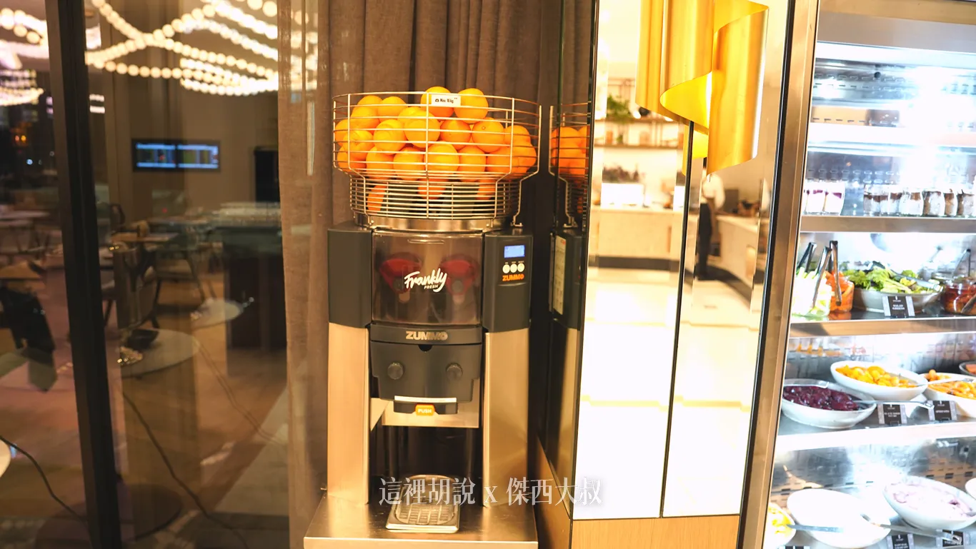 奧克蘭機場鉑爾曼飯店早餐區設置自動鮮榨果汁機，金屬果籃裝滿金黃柳橙，一旁冷藏櫃陳列著各式沙拉水果與甜點，提供傑西最喜歡的新鮮柳橙汁。
