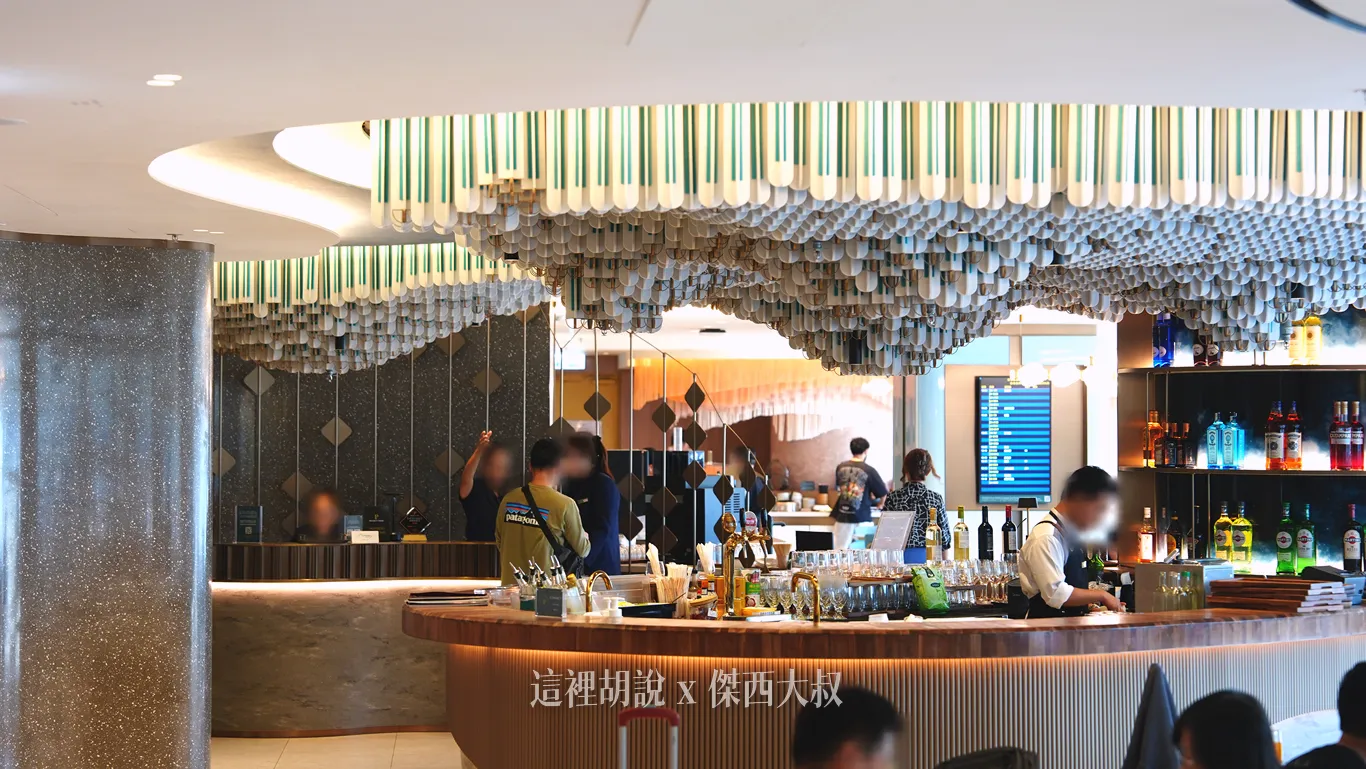 香港機場KYRA貴賓室天花板垂掛著波浪造型裝飾,左側為旅客報到的接待處理櫃檯,右側圓弧狀的調酒吧檯則陳列多款酒瓶與酒杯,營造明亮俐落的空間氛圍。 香港機場KYRA貴賓室天花板垂掛著波浪造型裝飾,左側為旅客報到的接待處理櫃檯,右側圓弧狀的調酒吧檯則陳列多款酒瓶與酒杯,營造明亮俐落的空間氛圍。