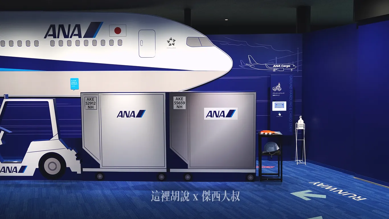 全日空訓練中心的體驗區以藍色展間為背景，展示印有全日空標誌的客機機身與拖車行李櫃拍照背板，一旁陳列地勤安全帽與背心供參觀者拍照留念。